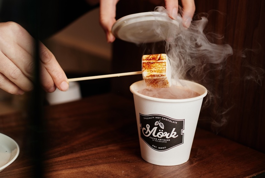 Marshmallow on a stick being held over a steaming takeaway cup, branded Mork.