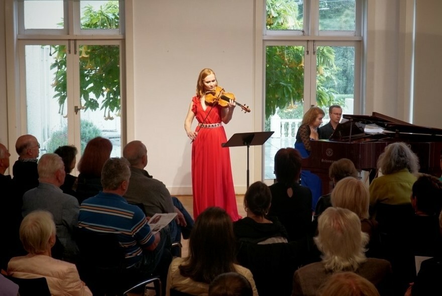 Woman in a red dress playing violin to a room of people.
