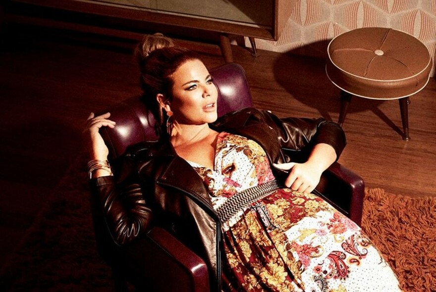 Female model reclining in a leather chair, wearing a leather jacket, metal belt and floral dress.