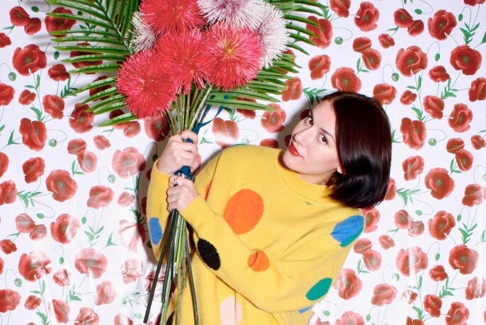 Model wearing yellow jumper with spots, holding flowers against rose-patterned wallpaper.