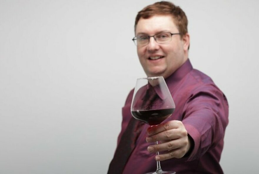 Man wearing a purple shirt and glasses, holding a glass of red wine at front with an extended arm.