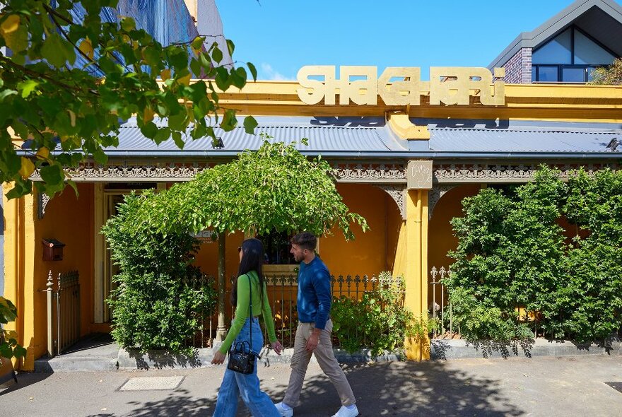 A couple walking past a yellow building surrounded by trees and a sign that reads 'Shakahari'.