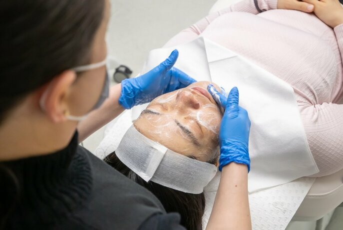 A customer lying on a bench being given a beauty treatment by a clinician wearing a mask and blue gloves.