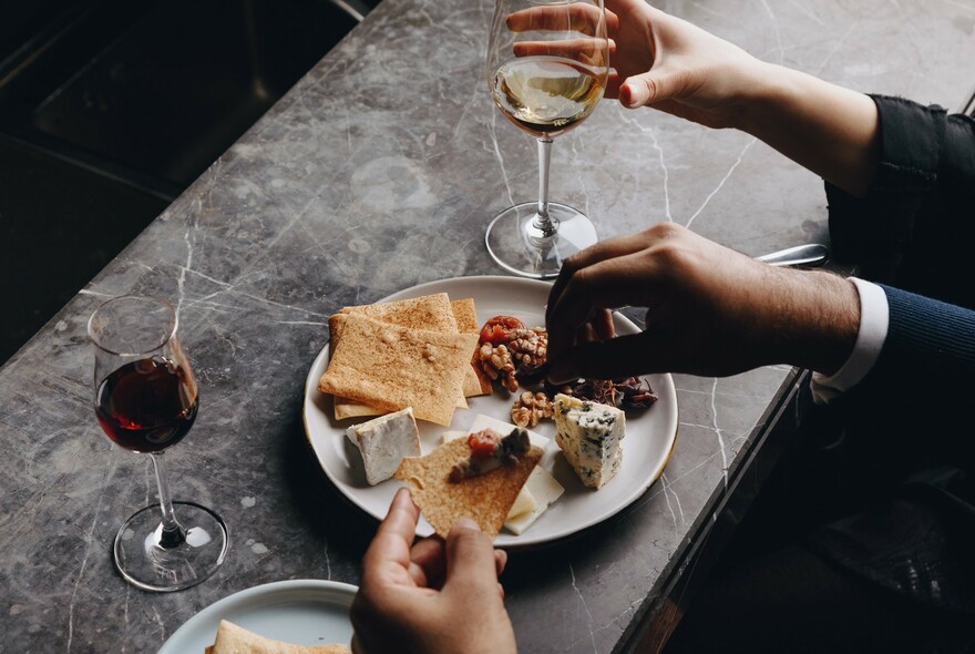 Diners' hands with glasses of wine and plate of cheese, crackers and charcuterie.