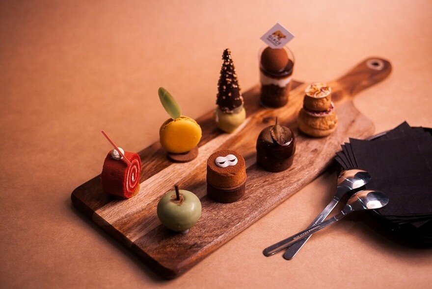 Wooden board full of small desserts, two small spoons resting on black serviettes to the right.
