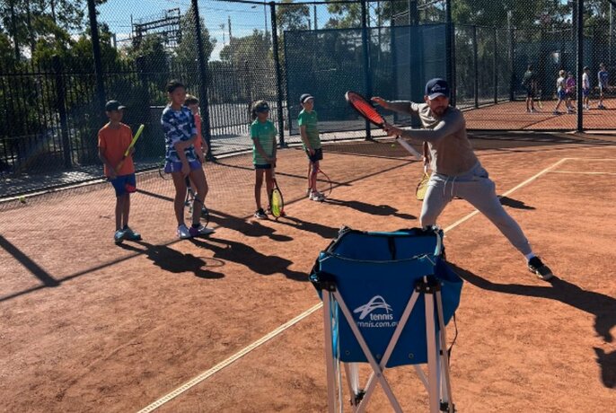 Adult tennis coach demonstrating a tennis pose and action to several children watching from the baseline.