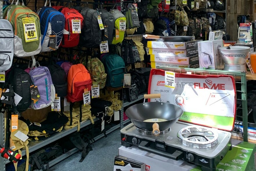 Interior of shop displaying racks of daypacks on the wall and a camping stove in the foreground.
