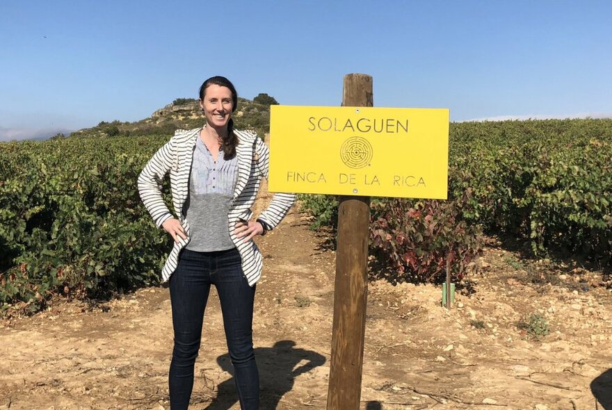 A woman smiling with her hands on her hips and standing outdoors in a vineyard next to a yellow sign with faded Spanish words written on it.