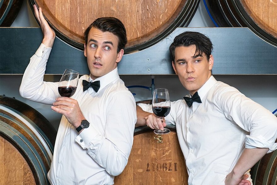 Comedians Sweeney Preston and Ethan Cavanagh, both wearing white shirts and black bow ties, each holding a glass of red wine and leaning against large oak wine barrels.