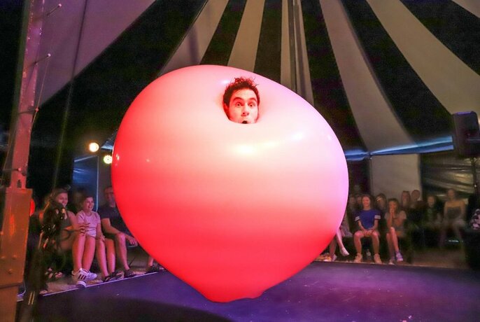 People in a circus tent watching a person's head pop out of a large pink balloon.