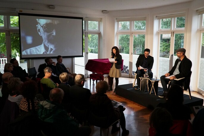 Three people on a small stage in a room, with an audience watching and a film screen mounted on the wall nearby.