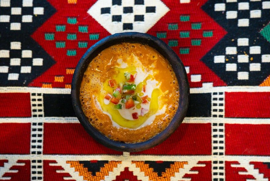 A dish of soup or stew presented on a tribal patterned cloth with red and blue geometric pattern.