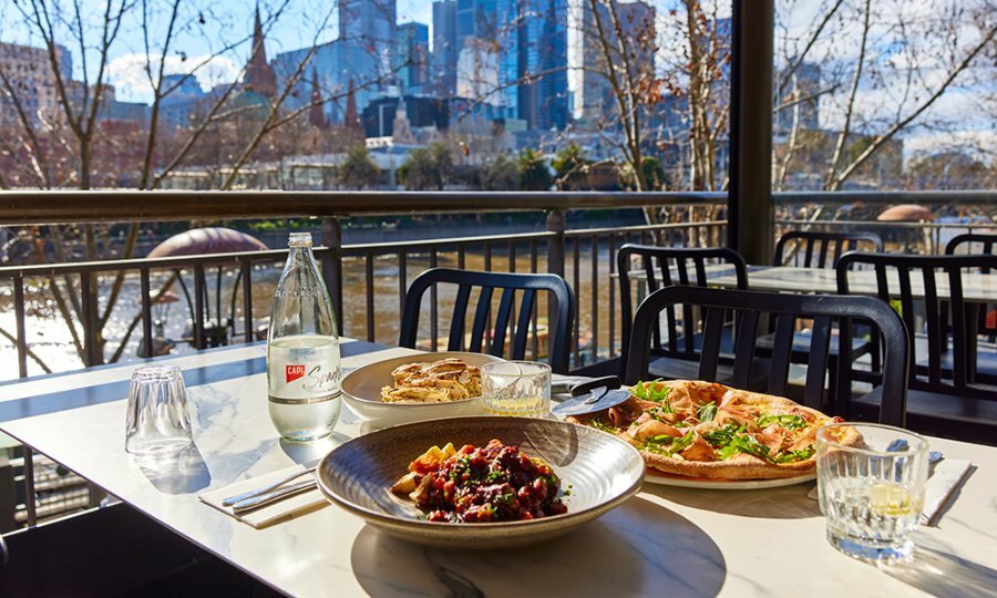 An outdoor restaurant by the river with pizza and pasta dishes on the table.