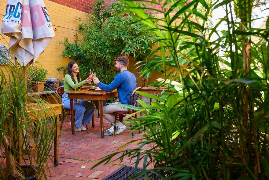 A couple sitting at a table in a leafy courtyard with drinks.