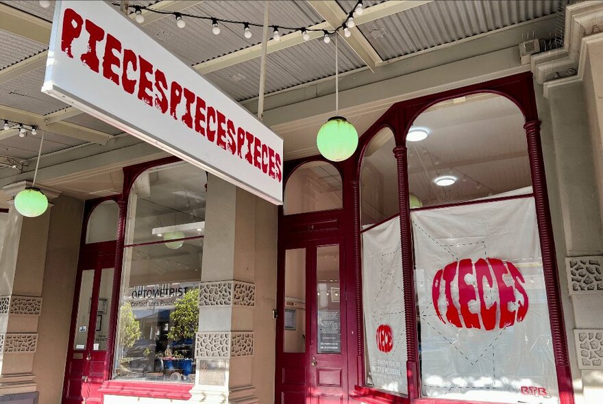 Exterior of a retail shop showing large windows, and a red door, and a hanging sign in front of the shop under a covered verandah that has the words PIECESPIECESPIECES written in red lettering.