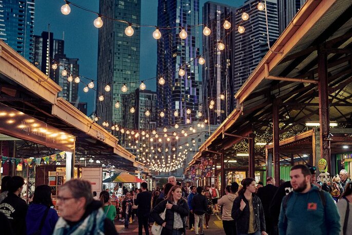 Shoppers under market awnings, with fairy lights above and cityscape at rear; night time.