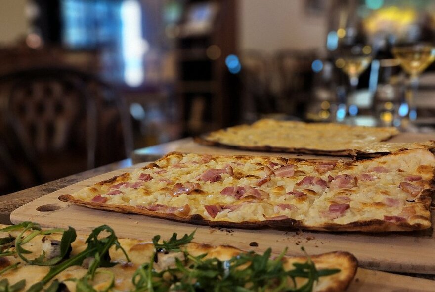 Large rectangles of pizza resting on wooden serving boards on a table, glasses of wine in the background.
