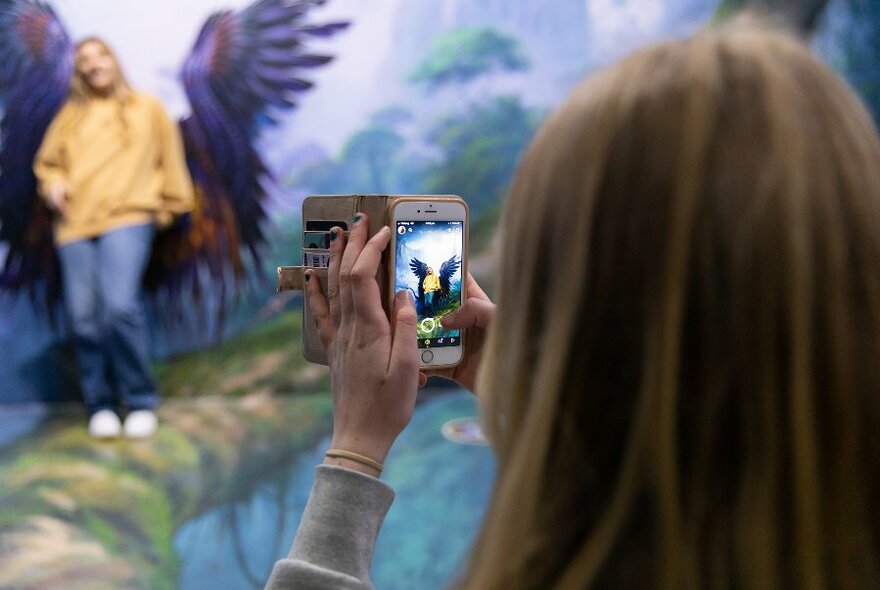 A person using their iPhone to film someone posing in front of a painting of wings.