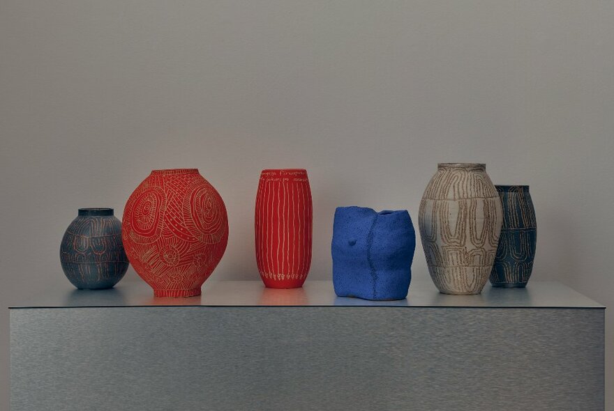 An image of six coloured vases or pots on an exhibition shelf. 