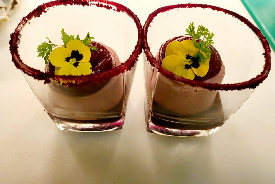 Food presented in two glasses, their rims rubbed with red powder and the creamy contents topped with yellow flowers.