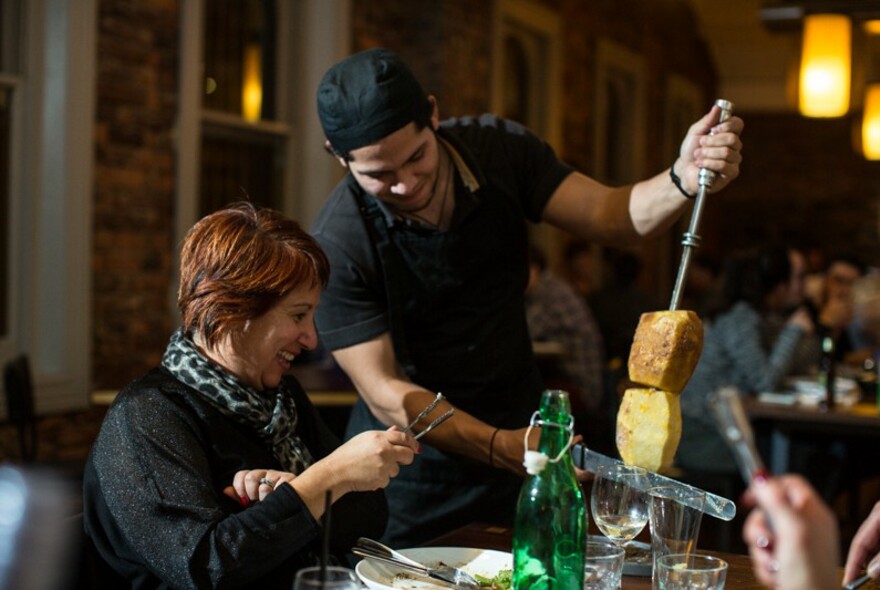 Waiter carving meat at a table with smiling customer with tongs.