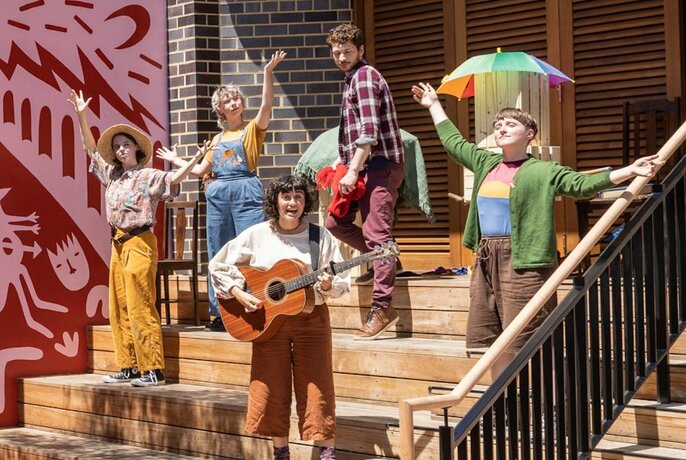 Five people on stairs, outdoors, one with guitar, another under rainbow umbrella, with with arms up in air. Pink and red mural to left.
