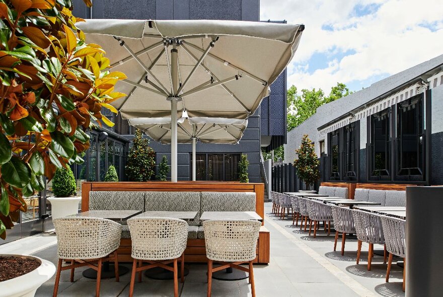 Outside dining area with banquette to rear, white chairs at front, large white umbrella overhead, similar set up along right hand side.