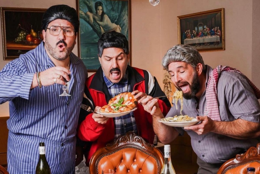 The trio of comedians of Sooshi Mango, in character costumes, eating food off plates they hold in their hands, one drinking a glass of red wine, in a lounge room like setting.