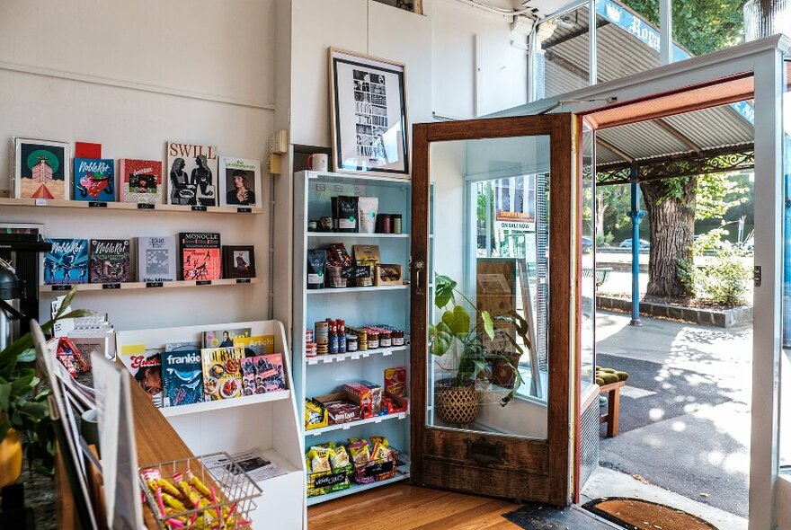 A milk bar with the door open. Shelves are filled with magazines and grocery items.