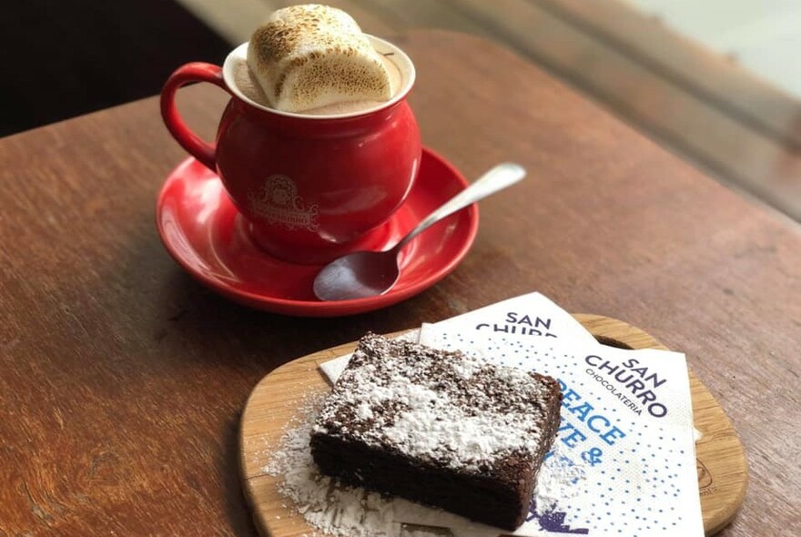 Red cup and saucer with hot chocolate and marshmallow, with icing sugar-dusted chocolate brownie on a wooden plate.