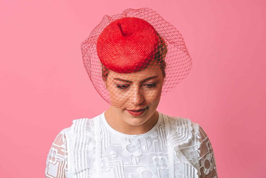 A model wearing a frilly white shirt and a red pillbox hat and veil, against a pink background.