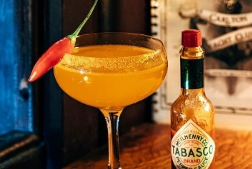 Orange coloured cocktail garnished with a small red chilli, a bottle of Tabasco next to it. 