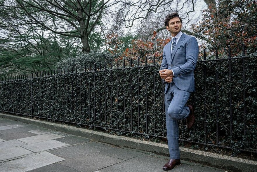 Male model in a grey suit, touching his cuffs and with one leg bent against park railings with hedge.