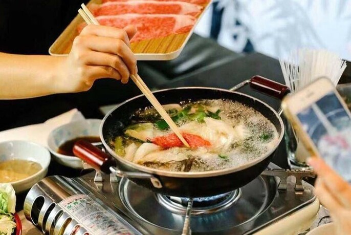 Hand using chopsticks to add meat to a wok heating on a tabletop grill.