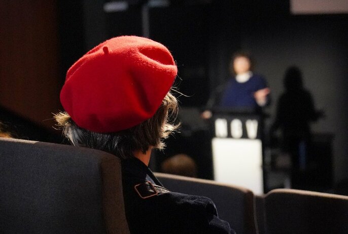 Looking over the shoulder of a person wearing a red beret to a performer on stage. 