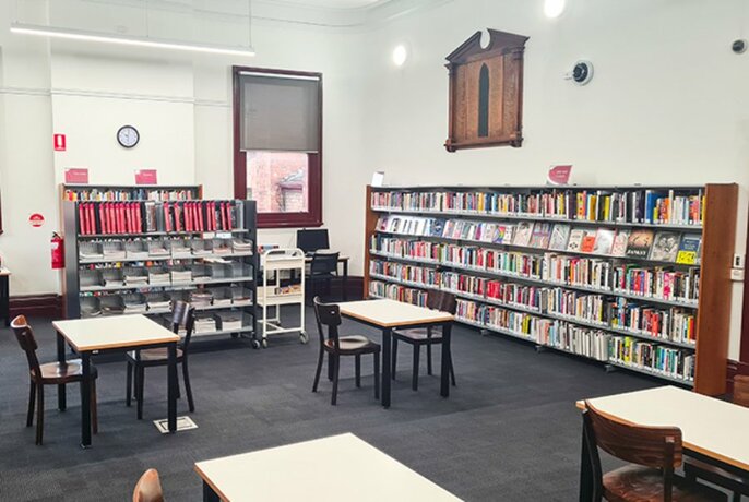 Small work tables and chairs, and shelves of books against the walls inside North Melbourne Library.