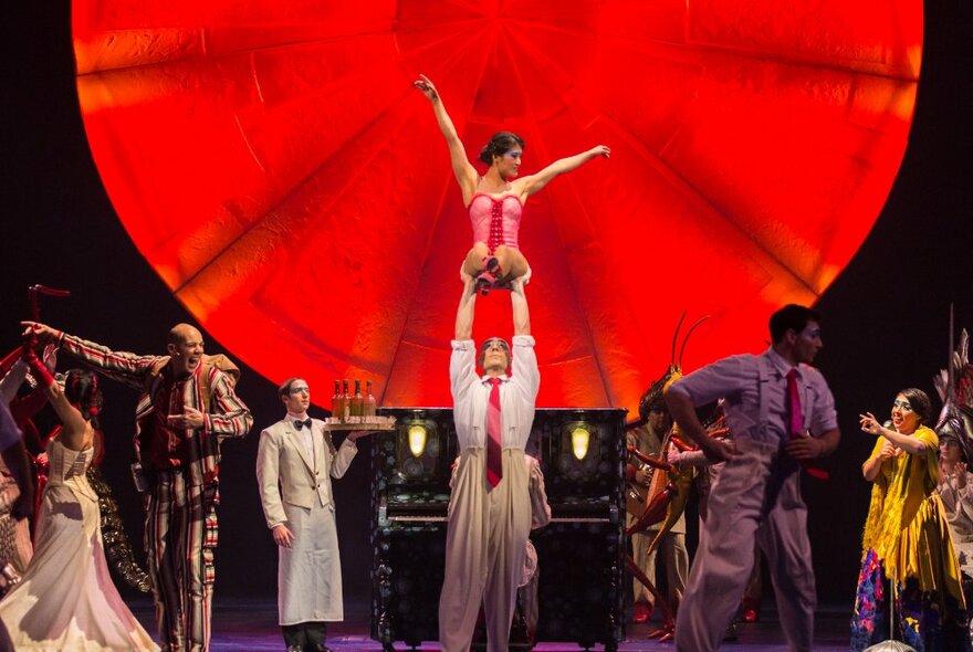 Performers on stage including acrobats, standing in front of a large round red theatre set.