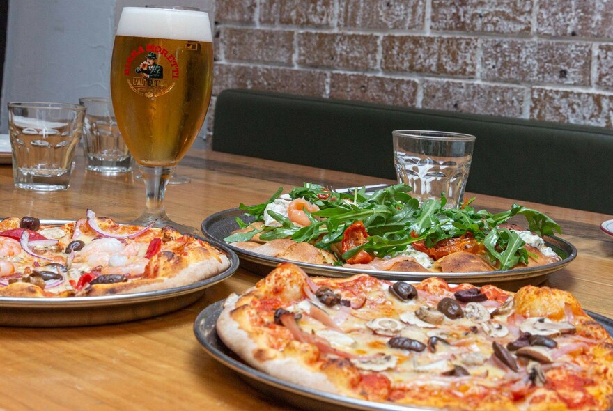 Table with three pizzas and glass of beer.