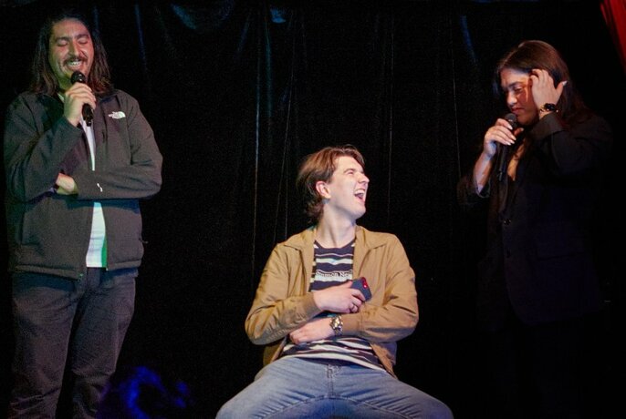 Three comedians performing on stage, one seated the other two holding microphones.