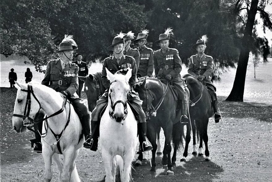 Men in military uniforms riding horses as part of a parade in a park.