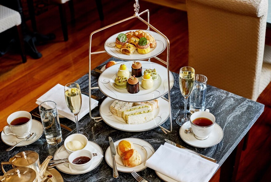 High tea selection of sweet and savoury food on three-tiered plates with cups of tea and coffee and glasses of champagne.