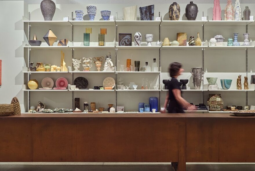 Blurred person in motion walking past a wall of shelving displaying art and craft objects, a wooden service counter in the foreground. 