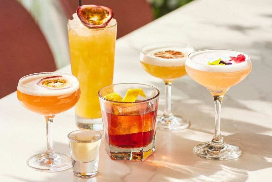 A lineup of summery drinks on a white table including cocktails and fruit punch with passion fruit.