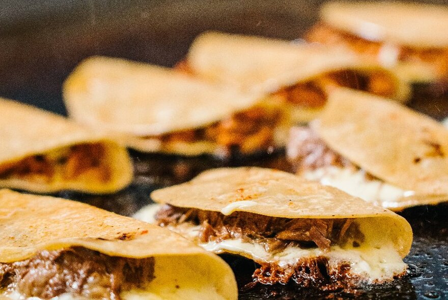 Several folded over tacos, filled with meat and cheese, sizzling on a grill hot plate.