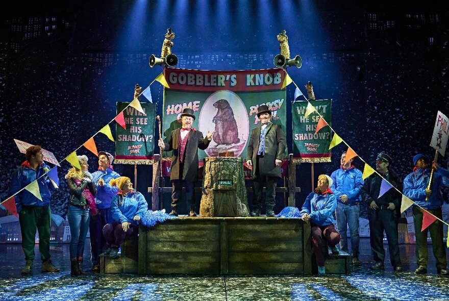 A theatre set-piece from the stage musical Groundhog Day with actors looking at two men in wearing suits and bowler hats standing in front of a sign.