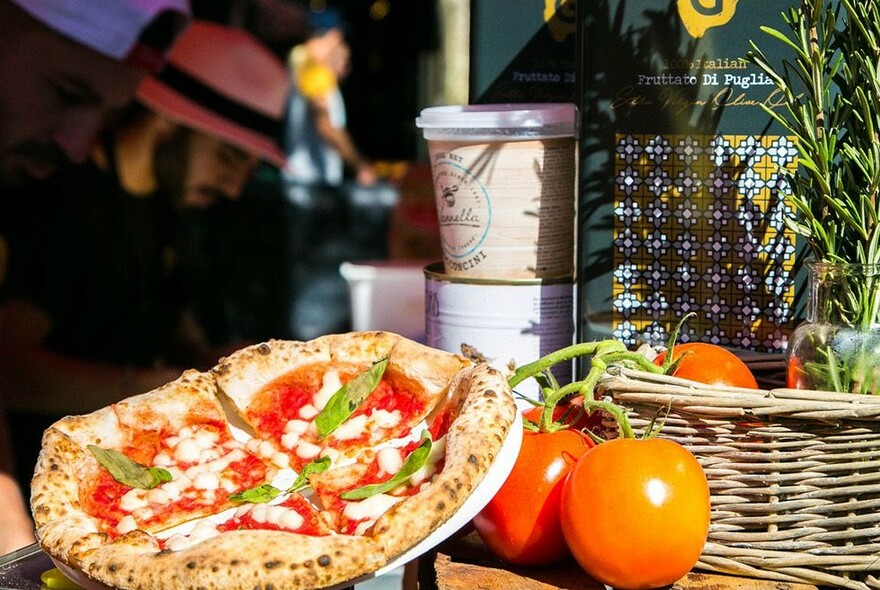 An authentic-looking margherita pizza in the sun next to a basket with ripe tomatoes and rosemary.