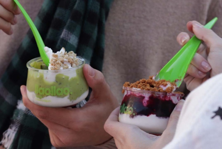 Green spoons being dipped into two small plastic cups with yoghurt and fruit.