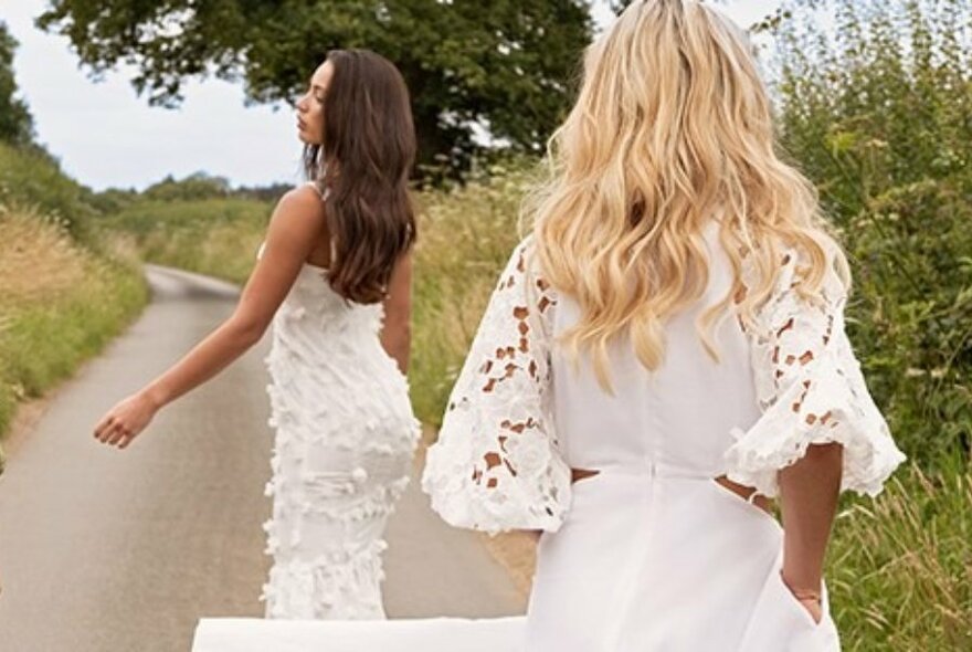 Models viewed from behind, wearing white dresses and walking along a country lane.