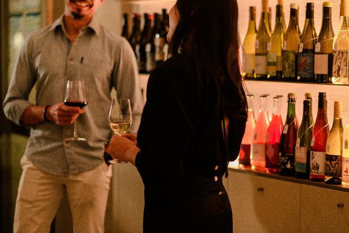 People holding glasses of wine next to illuminated shelves of wine.