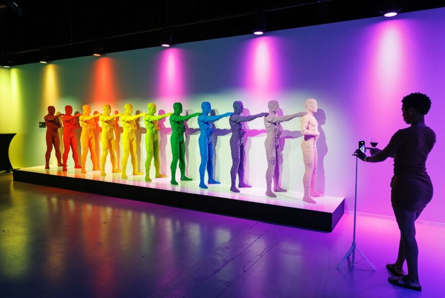 A row of 11 human sculptures, each one crafted from different coloured lego bricks, on display on a low platform in a large exhibition space, with a real person taking a photos of the display.